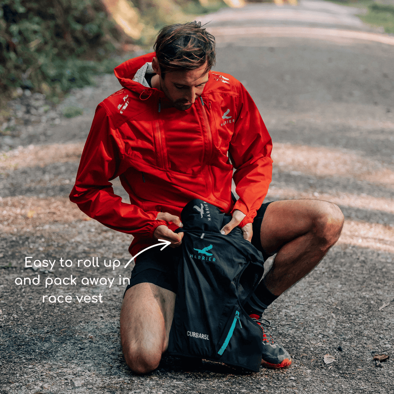 A man knelt down in a forest. He is packing away his waterproof trousers in to a running hydration vest.