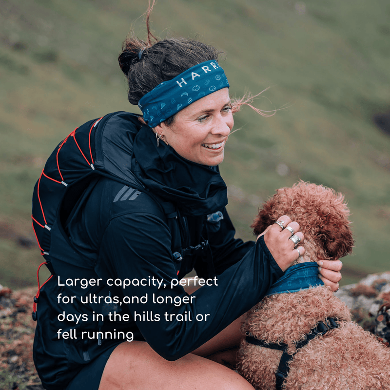 A woman wearing a running hydration vest, crouching next to a dog in the countryside.